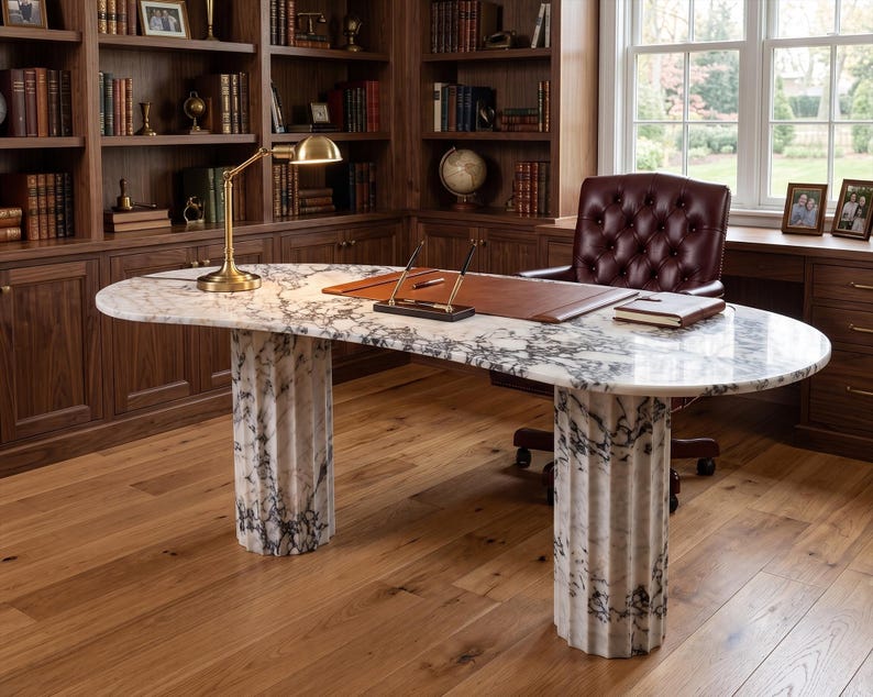 May include: An oval desk featuring a white and grey marble top supported by two fluted marble columns. The desk surface includes a brown leather desk pad, a gold-toned desk lamp, a pen stand with two pens, and a closed notebook.