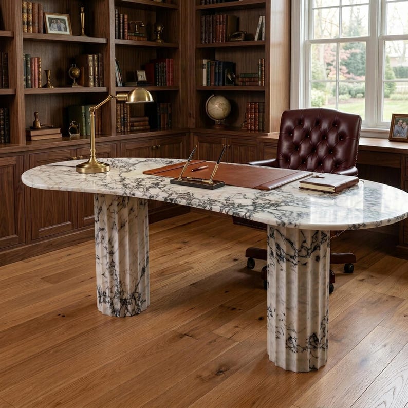 May include: An oval desk with a white and grey marble top supported by two fluted marble columns. The desk features a brown leather desk pad, a gold-toned lamp, a pen stand with two pens, and a closed notebook. A tufted burgundy leather chair sits behind.