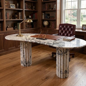 May include: An oval desk with a white and grey marble top supported by two fluted marble columns. The desk features a brass lamp, a brown leather desk pad with two pens, and a small notebook. A tufted burgundy leather office chair is positioned behind.