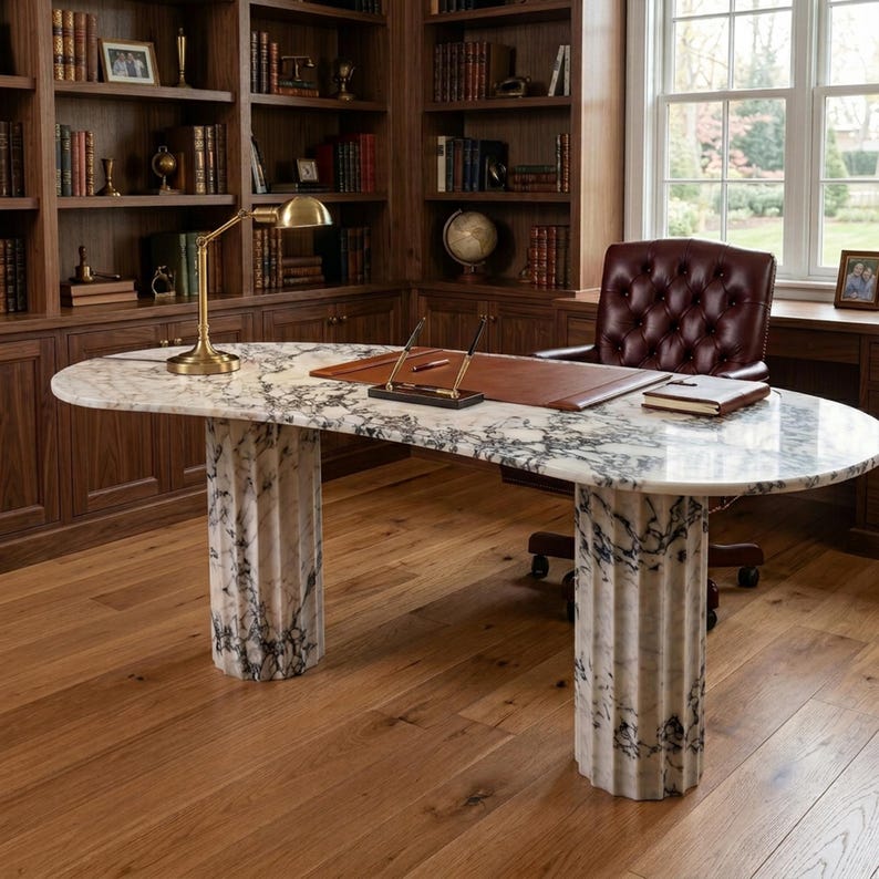 May include: An oval desk with a white and grey marble top supported by two fluted marble columns. The desk features a brass lamp, a brown leather desk pad with two pens, and a small notebook. A tufted burgundy leather office chair is positioned behind.