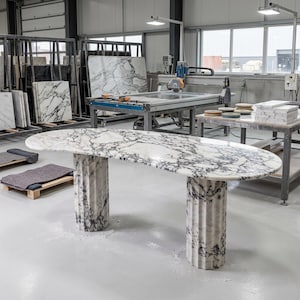 May include: An oval table crafted from white marble with prominent dark gray veining. The tabletop is supported by two cylindrical, fluted pedestals made of the same matching white and dark gray marble, standing on a smooth, light-colored floor.