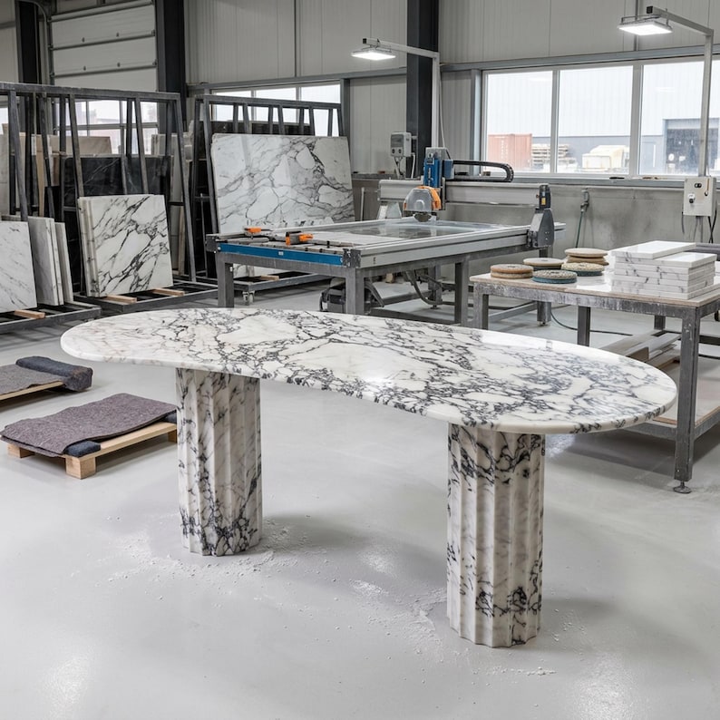 May include: An oval table crafted from white marble with prominent dark gray veining. The tabletop is supported by two cylindrical, fluted pedestals made of the same matching white and dark gray marble, standing on a smooth, light-colored floor.