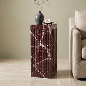 May include: A tall, rectangular pedestal table crafted from deep red marble with prominent white veining. The sides feature a vertical fluted design. On top sits a dark gray ceramic vase containing dried stems and a small white geode on a tray.