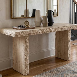 May include: A beige stone console table with a rough, natural edge and two thick slab legs. On top sits a rectangular wooden tray containing a small gold-toned compass and glasses, alongside three vases in varying heights and shades of cream and black.