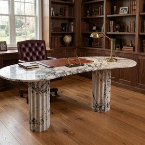 May include: An oval desk featuring a white marble top with grey veining supported by two fluted marble columns. The desk surface holds a brown leather desk pad, a black and gold pen set, and a brass desk lamp. A tufted burgundy leather chair sits behind.