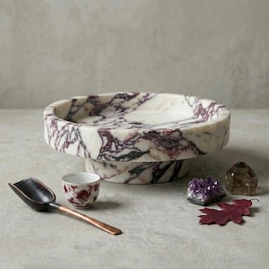 May include: A circular white marble pedestal bowl featuring prominent dark purple veining. Accompanying items include a small white ceramic cup with a red floral pattern, a dark metal scoop, a purple amethyst cluster, and a single dried red leaf.