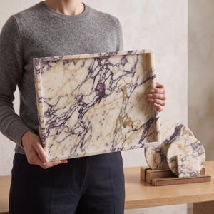 May include: A rectangular marble tray featuring a cream-colored base with prominent deep purple and gold veining. The tray has raised edges and a smooth, polished finish. Accompanying the tray are two circular marble coasters and a square coaster.