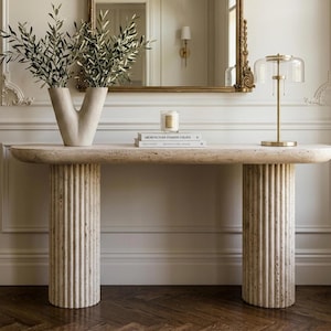 May include: A beige travertine console table with a smooth, elongated oval top supported by two thick, fluted cylindrical columns. The table surface holds a V-shaped vase with green branches, a small candle, two stacked books, and a brass lamp.
