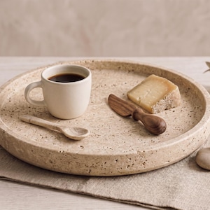 May include: A round, beige, speckled stone tray holding a small cream-colored ceramic cup filled with black coffee, a tiny stone spoon, a wooden cheese knife, and a wedge of hard cheese. The tray has a porous, natural texture throughout.