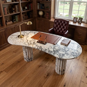 May include: An oval desk with a white and grey marble top supported by two fluted marble columns. The desk features a brown leather desk pad, a gold-toned metal desk lamp, a pen stand with two pens, and a small brown notebook placed on the surface.