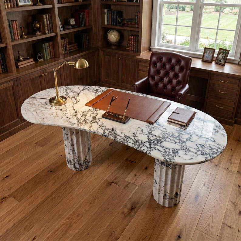 May include: An oval desk with a white and grey marble top supported by two fluted marble columns. The desk features a brown leather desk pad, a gold-toned metal desk lamp, a pen stand with two pens, and a small brown notebook placed on the surface.