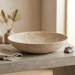 May include: A round, beige travertine stone vessel sink with a porous, natural texture. The basin features a wide, shallow bowl shape with a smooth interior rim and a distinctively pitted, organic surface finish throughout the exterior.