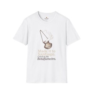May include: A white short-sleeved t-shirt featuring a graphic of a metallic Botafumeiro incense burner emitting smoke. Below the illustration, the text reads: Made it to Santiago. Cried at the Botafumeiro. The shirt has a round neckline and a label.