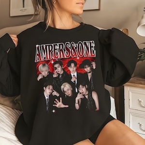 May include: A black crewneck sweatshirt featuring a graphic print of a group of seven individuals in dark clothing. Above the group, the text "AMPERS&ONE" is printed in a bold, red, distressed font against a red, lightning-like background.