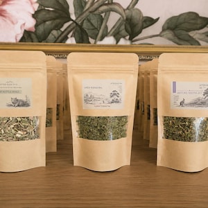 Puede incluir: Tres bolsas de papel kraft de pie que contienen té a granel. La etiqueta izquierda dice The Kettle Sings, la central Open Windows y la derecha Before Midnight. Cada bolsa tiene una ventana transparente que muestra las hojas de té secas.