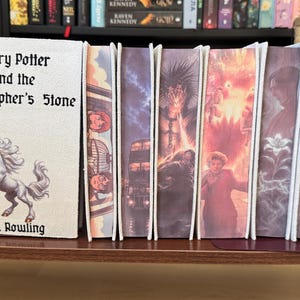 Könnte beinhalten: Ein Set aus sieben Harry-Potter-Büchern, die zwischen zwei dunklen Metallbuchstützen angeordnet sind. Das erste Buch hat einen weißen Einband mit einer Einhorn-Illustration und dem Text Harry Potter and the Philosopher's Stone von J.K. Rowling. Die restlichen sechs Bücher zeigen farbenfrohe, detaillierte Illustrationen auf ihren Buchschnitten.