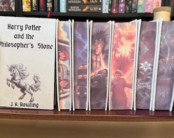 Könnte beinhalten: Ein Set aus sieben Harry-Potter-Büchern, die zwischen zwei dunklen Metallbuchstützen angeordnet sind. Das erste Buch hat einen weißen Einband mit einer Einhorn-Illustration und dem Text Harry Potter and the Philosopher's Stone von J.K. Rowling. Die restlichen sechs Bücher zeigen farbenfrohe, detaillierte Illustrationen auf ihren Buchschnitten.