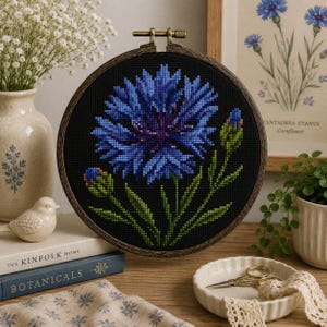 May include: A circular wooden embroidery hoop containing a cross-stitch pattern of a vibrant blue cornflower with green stems and leaves against a solid black background. The flower is depicted in full bloom with two smaller buds on the sides.