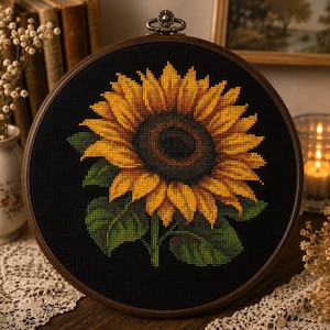 Puede incluir: Un bastidor de bordado circular que contiene un patrón de punto de cruz de un gran girasol con pétalos amarillos y naranjas vibrantes y un centro marrón oscuro, sobre un fondo de tela negra sólida, con hojas verdes en la base.