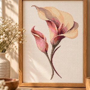 May include: A cross-stitch embroidery of two calla lilies in shades of cream, soft pink, and deep magenta, set against a white Aida cloth background. The artwork is encased in a light-toned wooden frame with a simple, clean-lined rectangular border.