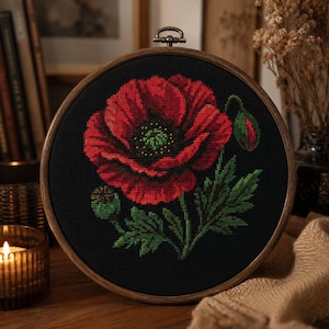 Può includere: Un telaio da ricamo circolare in legno contenente un disegno a punto croce di un grande papavero rosso vivace con un centro scuro, foglie verdi e due piccoli boccioli, su uno sfondo di tessuto nero con una vite di tensione in metallo.