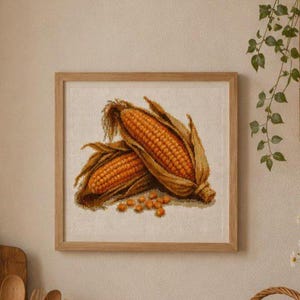 Könnte beinhalten: Ein gerahmtes Kreuzstichbild, das zwei goldgelbe Maiskolben mit getrockneten braunen Hüllblättern und einige lose Körner am unteren Rand zeigt. Das Kunstwerk befindet sich in einem hellen Holzrahmen mit quadratischem Profil und klaren Kanten.