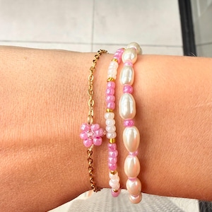 Op de afbeelding: Een set van drie armbanden bestaande uit een goudkleurige metalen ketting met een roze kralen bloemhanger, een streng van afwisselende roze en witte kraaltjes met gouden tussenstukjes, en een streng van witte ovale parels met roze kraaltjes.