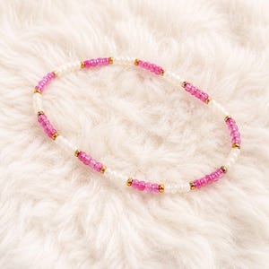 Op de afbeelding: Een kralenarmband met een herhalend patroon van kleine, doorschijnende roze kralen en ondoorzichtige witte kralen, gescheiden door kleine, reflecterende goudkleurige metalen tussenkralen, gerangschikt in een cirkelvormige lus op een witte ondergrond.