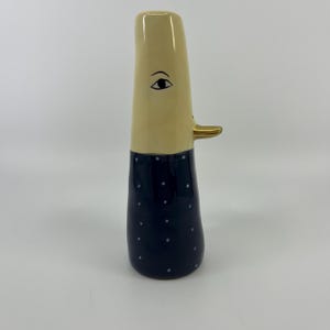 Puede incluir: Un jarrón de cerámica alto y esbelto con una sección superior de color crema que presenta un solo ojo negro y un pequeño pico dorado que sobresale. La sección inferior es de color azul oscuro con un patrón de pequeños puntos blancos.