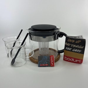 May include: A clear glass teapot with a black lid and handle, a central infuser, and a cork base. Two clear glass mugs with black spoons are included. A product tag reads "GIVE UP BAD DESIGN FOR GOOD" and "bodum" with a small instruction card.