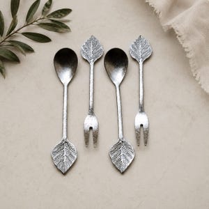 May include: A set of four silver-toned metal utensils comprising two spoons and two two-pronged forks. Each utensil features a textured leaf-shaped handle and a matching leaf-shaped detail at the base of the stem, with a weathered metallic finish.