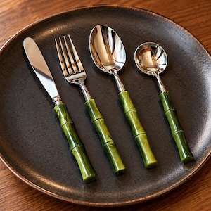May include: A four-piece stainless steel flatware set consisting of a dinner knife, a dinner fork, a dinner spoon, and a smaller teaspoon. Each utensil features a polished silver finish and a handle designed to resemble green bamboo stalks.