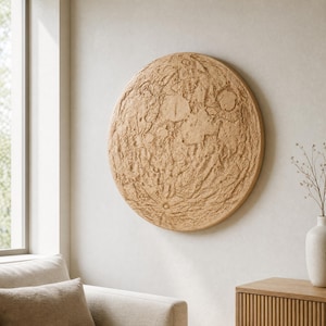 Puede incluir: Una pieza de arte mural circular que presenta un relieve detallado y texturizado de la superficie lunar en un color arena claro. La obra muestra cráteres, crestas y llanuras lunares con una apariencia tridimensional realista montada en la pared.