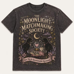 Puede incluir: Una camiseta gris carbón con lavado ácido que presenta un estampado gráfico de dos gatos negros, una bola de cristal con un corazón, una taza de té y cartas del tarot. El texto dice THE MOONLIGHT MATCHMAKING SOCIETY y Fate, Tea & Questionable Decisions.