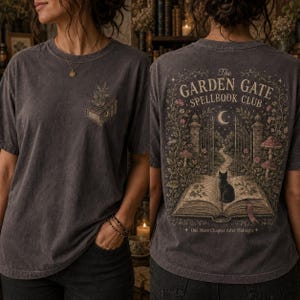 Puede incluir: Una camiseta gris carbón con un pequeño gráfico de un libro en el pecho. La parte posterior muestra una gran ilustración de un libro abierto con un gato negro, una puerta de jardín, setas y el texto The Garden Gate Spellbook Club.