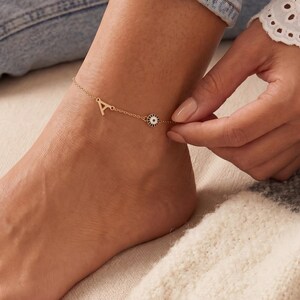 Puede incluir: Una delicada tobillera de cadena en tono dorado que presenta un pequeño dije con la letra A y un dije circular de ojo turco con detalles en azul, blanco y negro. La tobillera está sujeta alrededor del tobillo con una fina cadena de eslabones.