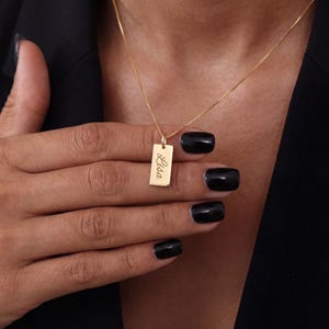 Puede incluir: Un collar de tono dorado que presenta un pequeño colgante rectangular con el nombre Lisa grabado en letra cursiva. El colgante cuelga de una cadena fina y delicada de tono dorado, creando una pieza de joyería sencilla y refinada.