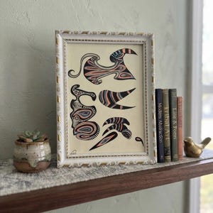 May include: A framed artwork featuring abstract, organic shapes in shades of black, peach, and blue on a cream background. The white frame has gold accents. Beside it are three books titled Modern Abstraction, Tribal Art, and Line & Form.