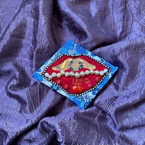 May include: A diamond-shaped fabric ornament featuring a red center panel decorated with a metallic watch component, a row of white pearls, small red beads, and a single orange striped bead, all bordered by a blue and white floral fabric.