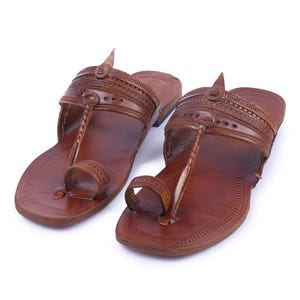 May include: A pair of brown leather sandals featuring a wide strap across the top with intricate embossed patterns, a central braided leather strip, and a toe loop. The flat soles have a rounded shape with decorative stitching along the edges.