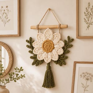 May include: A wall hanging featuring a crocheted daisy with white petals and a yellow center, accented by green crocheted leaves and a green tassel. The piece is suspended from a light-colored wooden dowel by a natural fiber cord.