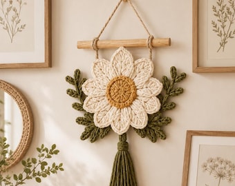 May include: A wall hanging featuring a crocheted daisy with white petals and a yellow center, accented by green crocheted leaves and a green tassel. The piece is suspended from a light-colored wooden dowel by a natural fiber cord.