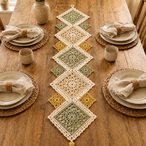 May include: A long, rectangular crochet table runner featuring a geometric pattern of interconnected squares in cream, sage green, and mustard yellow. The runner is finished with a decorative tassel at the bottom end and intricate lace-like stitching.