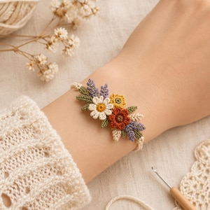 Puede incluir: Una pulsera de macramé con un arreglo central de flores de ganchillo en blanco, amarillo y naranja óxido, acentuado con hojas verdes y ramitas moradas. La pulsera está hecha de un cordón fino de color crema atado alrededor de la muñeca.