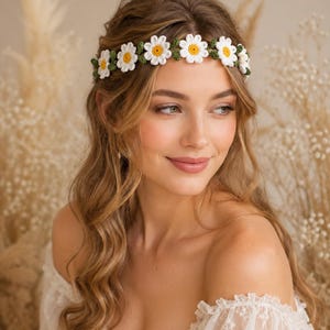 May include: A crochet headband featuring a series of white daisy flowers with yellow centers, connected by small green leaves. The floral band is worn across the forehead, showcasing detailed white petals and green foliage crafted from yarn.