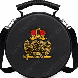 Op de afbeelding: Een ronde zwarte tas met een handvat aan de bovenkant en een verstelbare schouderriem. De voorkant is voorzien van een geborduurd embleem van een tweekoppige adelaar met een rode kroon, een gouden driehoek met het getal 33 en een zwaard.