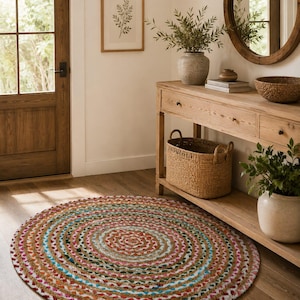 Puede incluir: Una alfombra redonda trenzada que presenta un patrón concéntrico de tiras de tela multicolor en tonos rojo, azul, verde, amarillo y blanco, tejidas junto con fibras de yute de color tostado natural para crear un revestimiento de suelo circular.