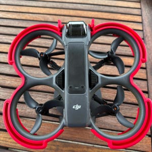 May include: A dark gray DJI drone featuring four integrated propeller guards with bright red outer rims. The central body displays the DJI logo, a camera lens at the front, and four black propellers positioned within the protective circular frames.