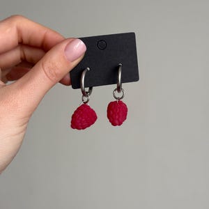 Peut inclure: Une paire de boucles d'oreilles créoles avec des breloques pendantes en forme de framboises texturées rose vif. Les boucles d'oreilles sont fixées sur une carte de présentation rectangulaire noire avec un trou circulaire, en métal argenté.
