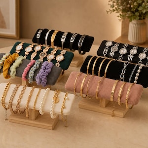 May include: A collection of multi-tiered jewelry display stands featuring velvet-covered bars in black, dark green, gray, white, and dusty pink. These stands hold various gold and silver-toned bracelets, wristwatches, and colorful fabric hair scrunchies.